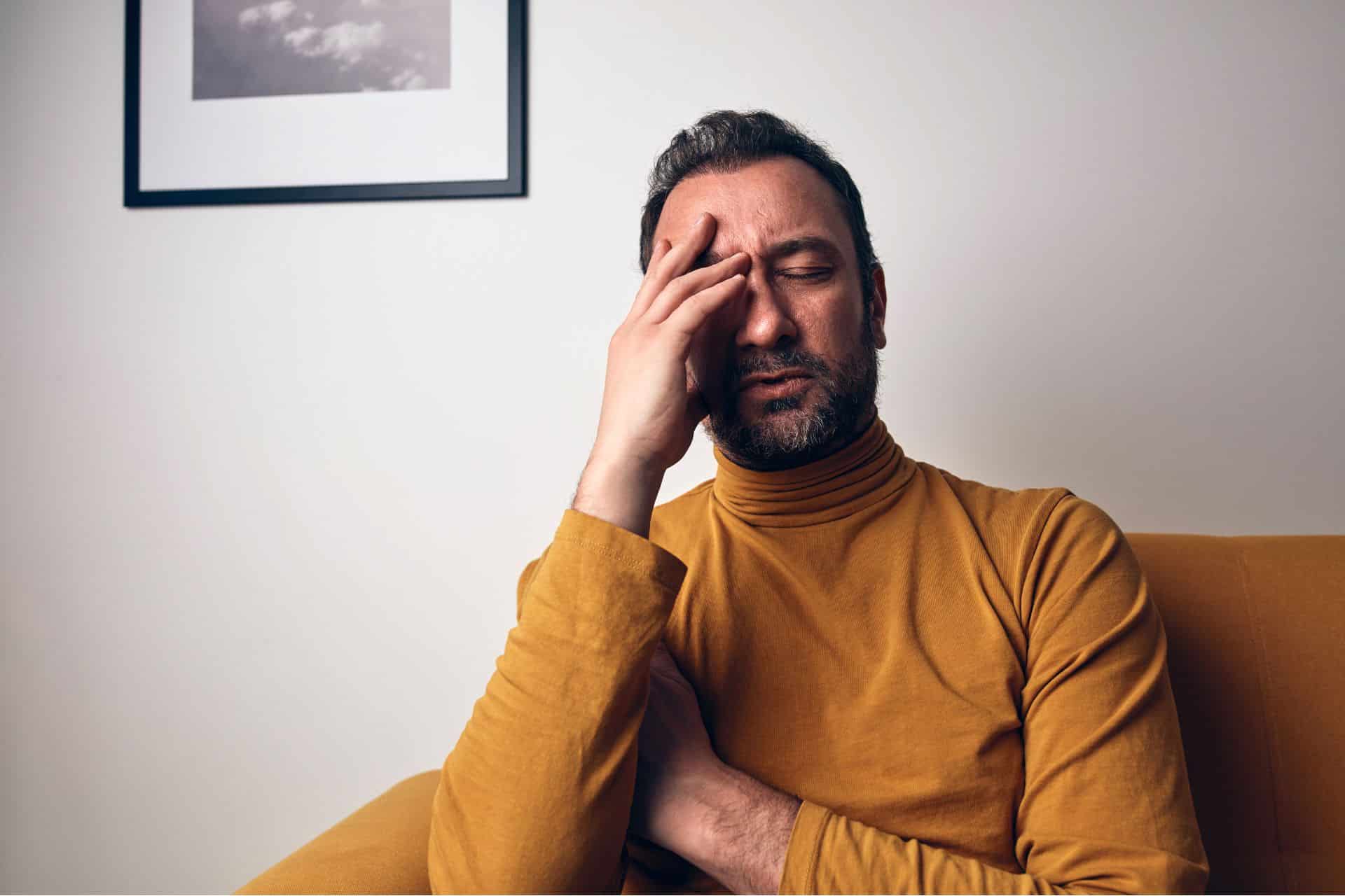 A man in a mustard turtleneck sits on a sofa with eyes closed and hand on his forehead, clearly distressed—an emotional moment that highlights the need for supportive practices like a mantra for anxiety.