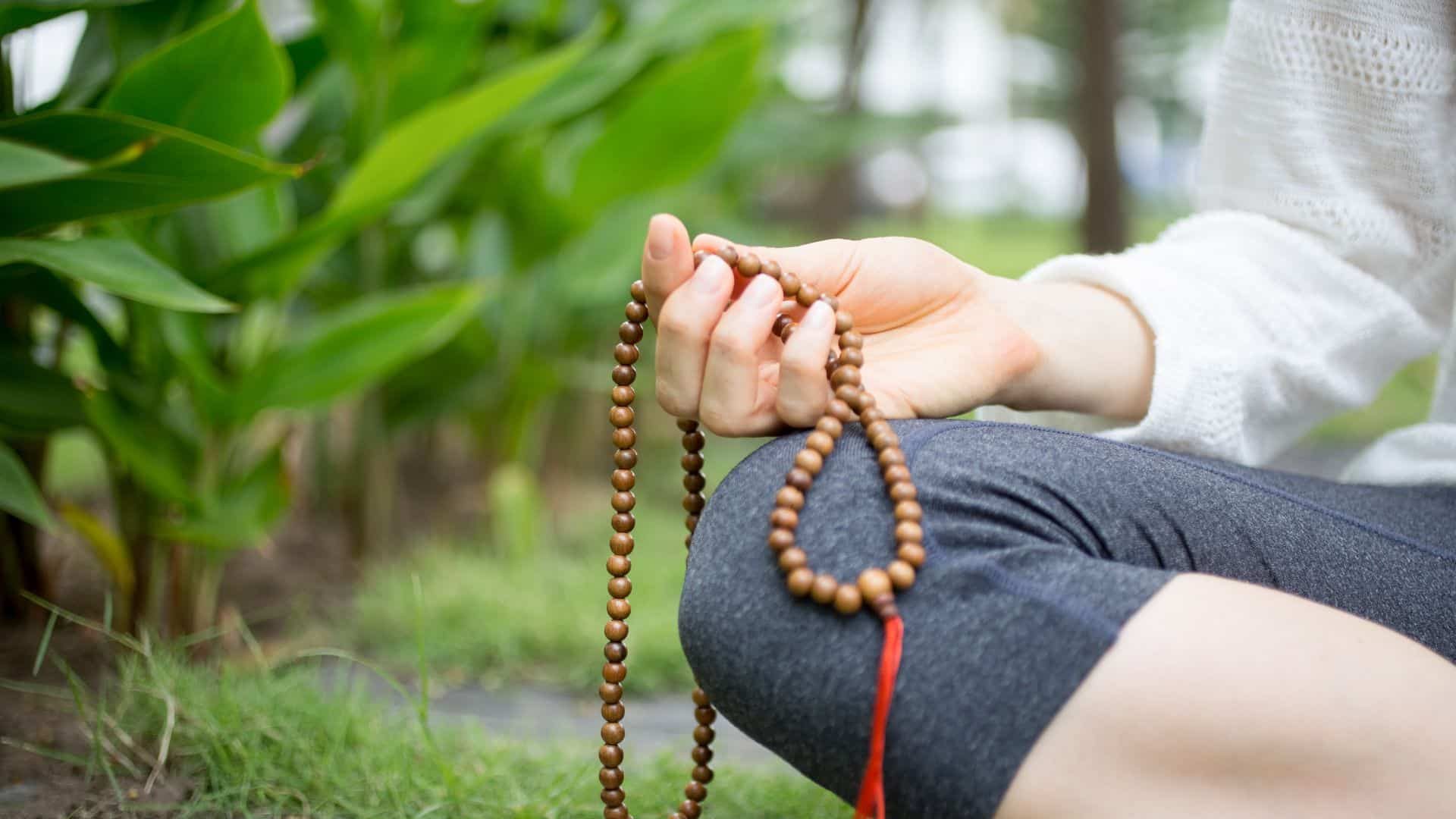 Hand Holding Mala Beads Outdoors A close-up of a person seated in meditation, gently holding mala beads in their hand in a peaceful garden setting—symbolizing the grounding use of a mantra for anxiety.